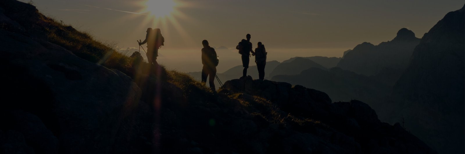 Silhuetas de três pessoas em destaque durante uma trilha na montanha ao pôr do sol, com picos de montanhas ao fundo, representando a jornada de autodescoberta, mesmo com os desafios.