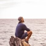 A man sitting on rocks by the ocean, reflecting under the warm sunrise.